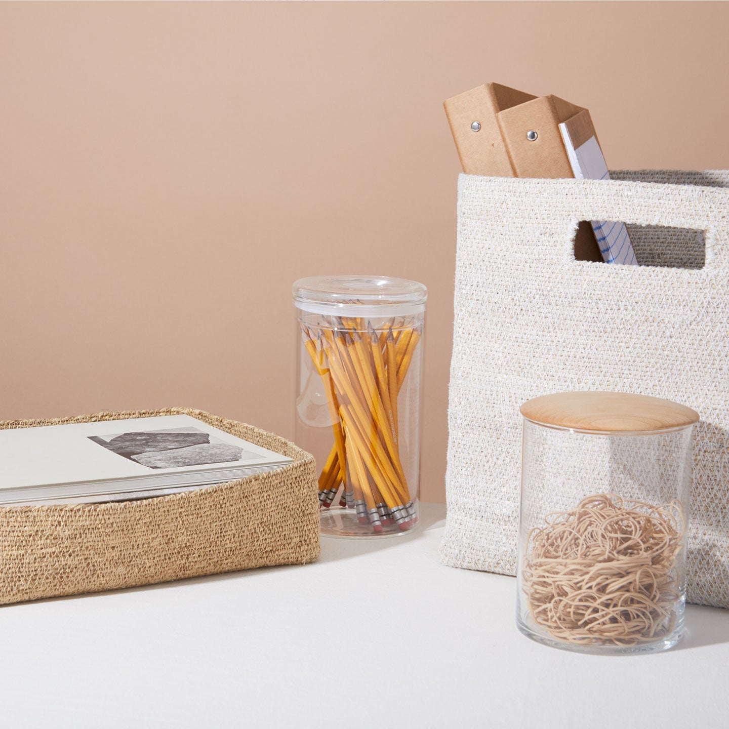 Assorted office supplies including a woven basket with papers, a clear jar with pencils, a bag with binders, and a glass jar filled with rubber bands, arranged on a white surface.