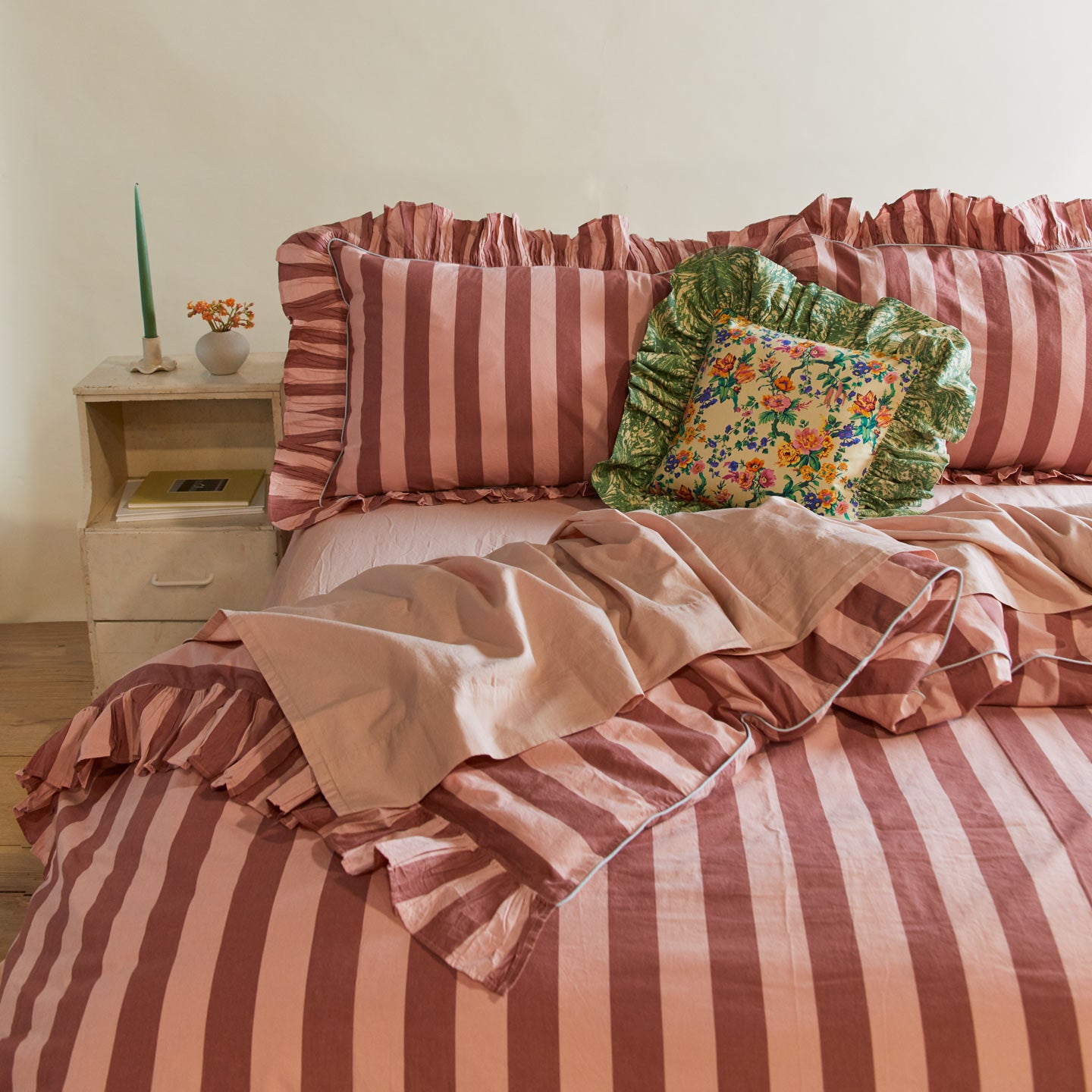 Messy bed with ruffled duvet cover and pillowcases with ruffled floral throw pillow. Off to the side is a green candlestick in a ceramic holder and florals in a vase.