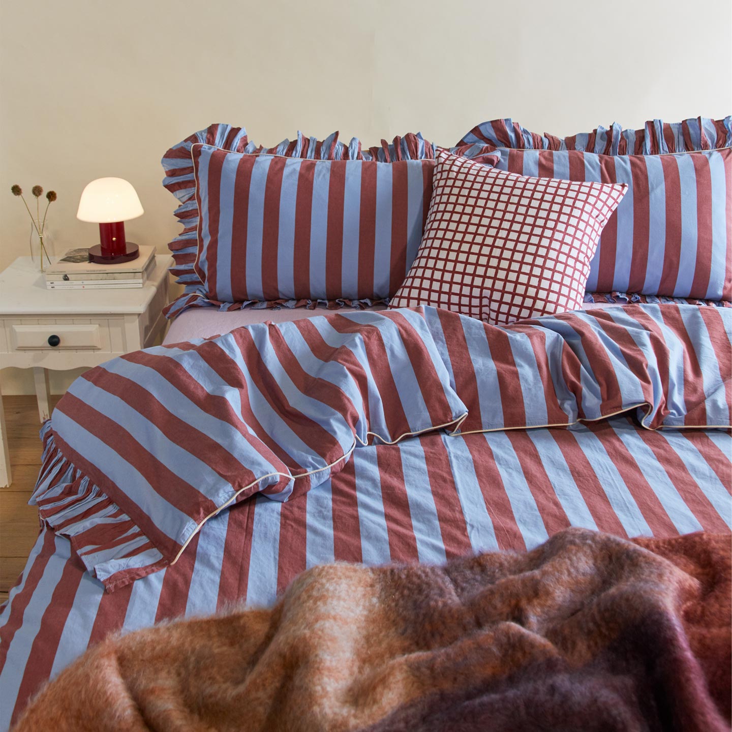 Messy bed with ruffled duvet cover and pillowcases with a mohair throw and checked throw pillow. Off to the side is a red lamp and florals in a glass vase.